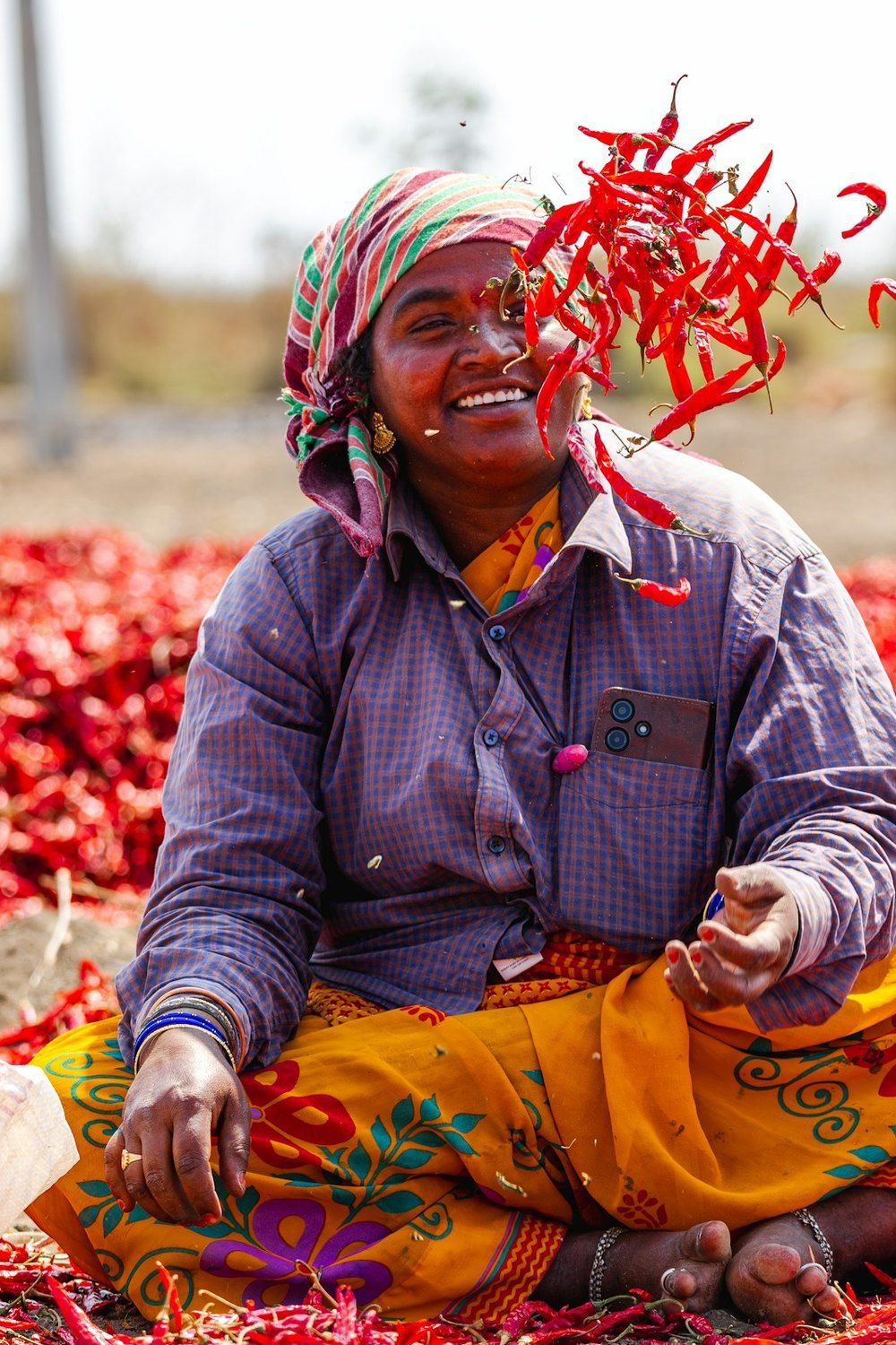 A smiling woman sits among red chillies drying in the sun, tossing a handful into the air